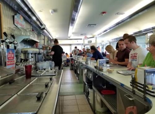 A busy diner interior with patrons seated at the counter and tables, and a server preparing food in the background.