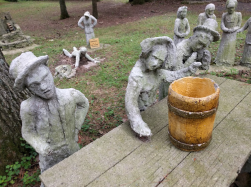 A group of weathered sculptures in a park, featuring figures around a wooden barrel on a stone table.