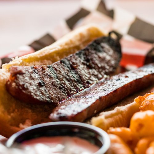 A close-up of a grilled sausage on a bun, accompanied by crispy snacks and dipping sauce on a wooden surface.