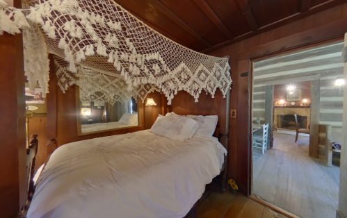 Cozy wooden bedroom with a lace canopy, mirror, and a view of a rustic living area with a fireplace.
