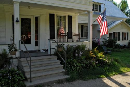 A charming porch with rocking chairs, an American flag, and lush greenery in front of a white house.