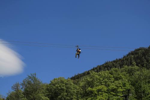 A person zip-lining through a clear blue sky, surrounded by green trees and a mountainous backdrop.