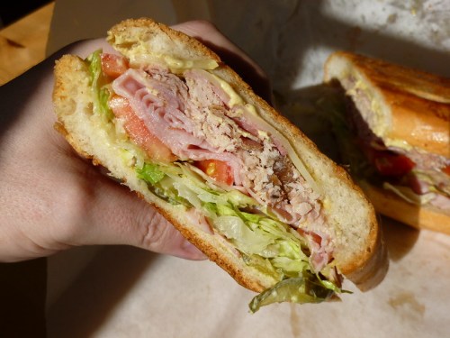 A hand holding a cross-section of a sandwich filled with lettuce, tomato, ham, and other ingredients on toasted bread.