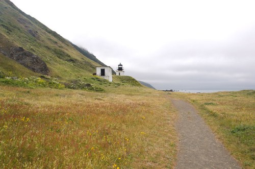 A winding path leads to a lighthouse on a grassy hillside, with a cloudy sky and ocean in the background.