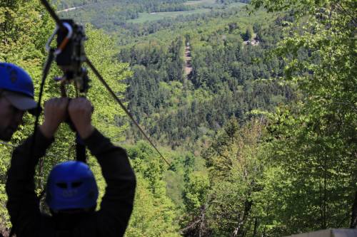 Two people in helmets prepare to zip line over a lush green landscape with trees and hills in the background.