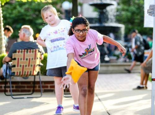 A girl in a pink shirt throws a yellow bean bag while another girl watches in a park setting with a fountain.