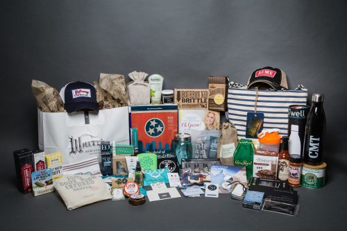A collection of various gift items, including snacks, drinks, and branded merchandise, arranged on a gray background.