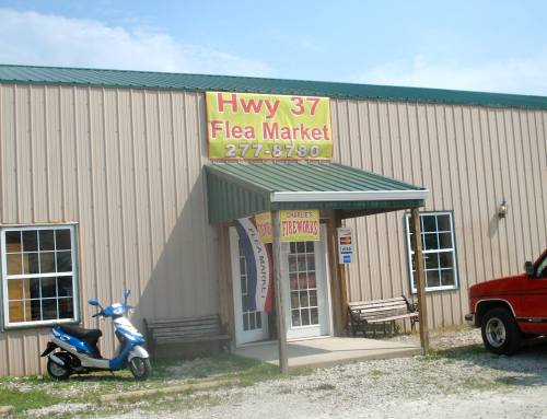 Exterior of Hwy 37 Flea Market building with a sign, benches, a blue scooter, and a red truck parked outside.