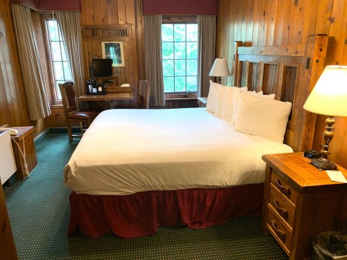 Cozy wooden hotel room featuring a queen bed, desk, and window with natural light. Warm, rustic decor throughout.
