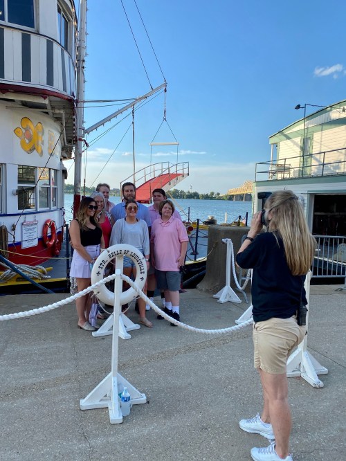 A group of people poses for a photo near a boat, with a scenic waterfront and buildings in the background.