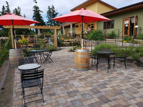 Outdoor patio with black tables and chairs, red umbrellas, and a garden area near a building with green and yellow walls.