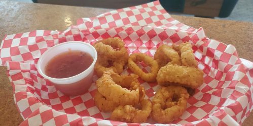 A basket of crispy fried calamari rings with a side of marinara sauce on a red and white checkered paper.