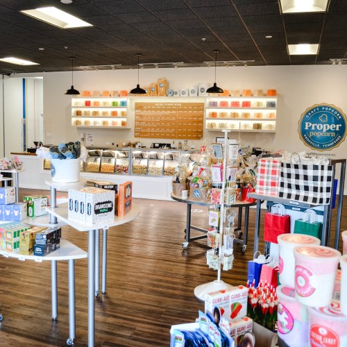A bright popcorn shop interior with various popcorn flavors, snacks, and colorful packaging displayed on shelves.