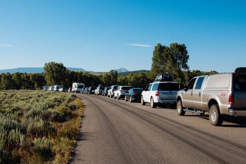 A long line of vehicles parked along a road, surrounded by trees and mountains under a clear blue sky.