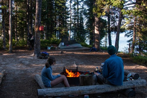 A campsite in the woods with a campfire, a tent, and two people enjoying the outdoors by a lake.