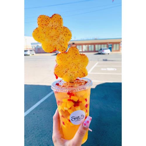 A colorful drink topped with flower-shaped pineapple slices and sprinkled with chili, held in a hand against a sunny background.