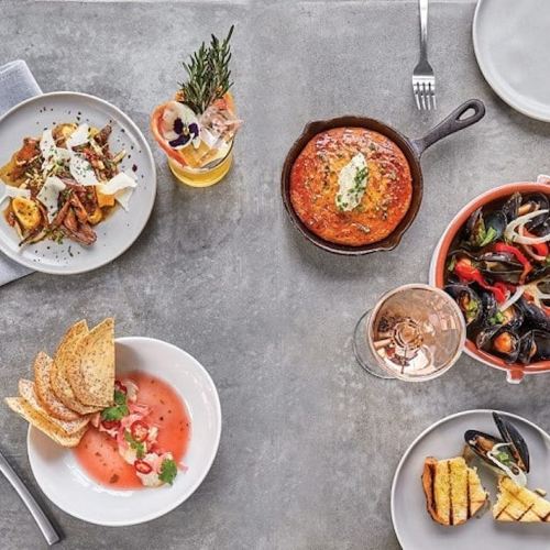 A table spread with various dishes: pasta, seafood, a baked dish, toast, and drinks, all elegantly presented.