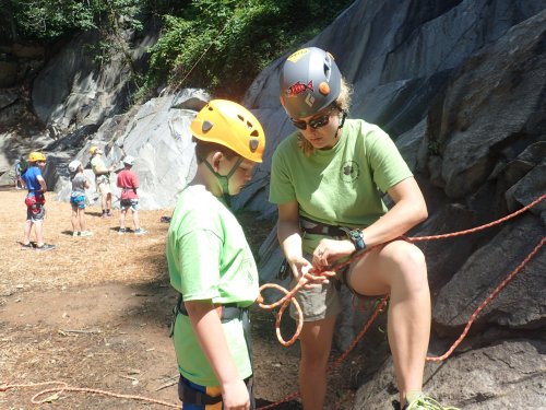 A climbing instructor teaches a child how to tie a knot, with other climbers in the background at a rock climbing site.
