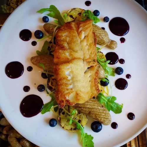 A beautifully plated dish featuring fried fish on a bed of grilled vegetables, garnished with greens and berry sauce.