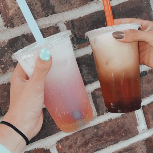 Two hands holding colorful drinks in clear cups with straws, against a brick wall background.