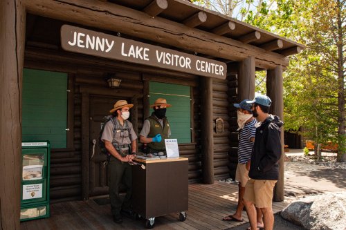 A park ranger speaks with visitors outside the Jenny Lake Visitor Center, surrounded by trees and a log cabin structure.