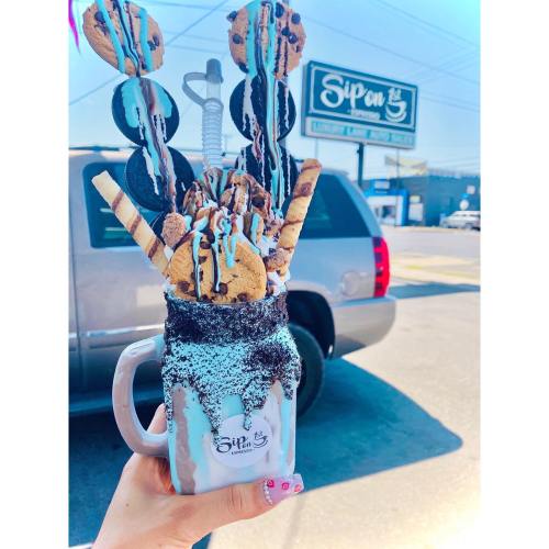 A hand holds a large, colorful milkshake topped with cookies, chocolate, and sprinkles, with a shop sign in the background.