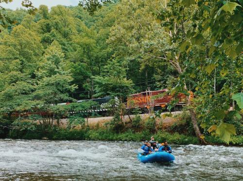 A group of people in a blue raft navigating a river, with lush green trees and a train passing in the background.
