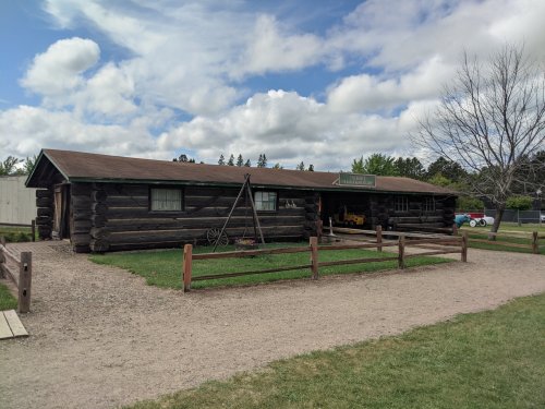 A rustic log cabin with a grassy area and wooden fence, set against a cloudy sky.