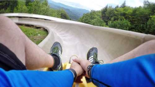 A person's legs on a luge track, holding hands with another person, with mountains in the background.