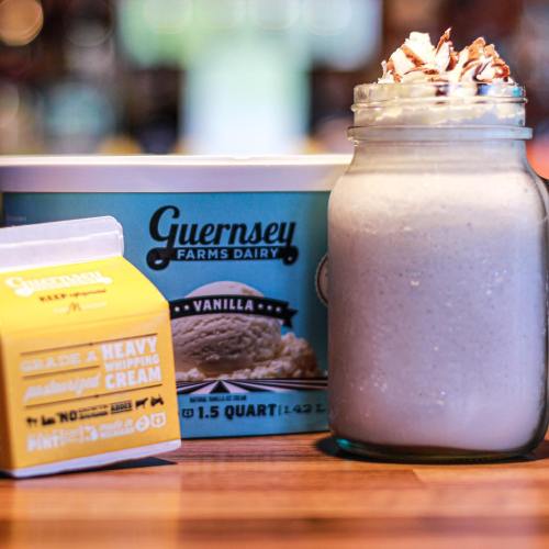 A jar of vanilla milkshake topped with whipped cream, next to a carton of heavy whipping cream and ice cream container.