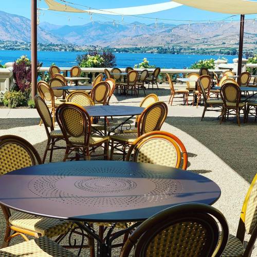 Outdoor dining area with empty tables and chairs overlooking a lake and mountains, under a sunny sky.