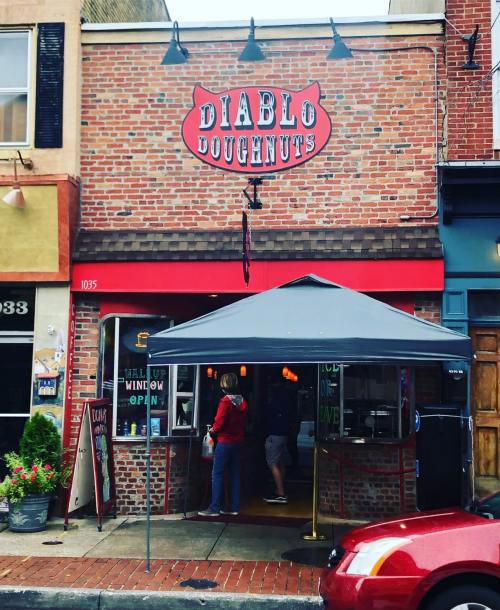 A brick storefront with a red sign reading "Diablo Doughnuts," featuring a canopy and a customer at the window.