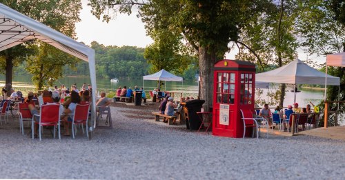 A lakeside gathering with people seated at tables under tents, featuring a red phone booth nearby.