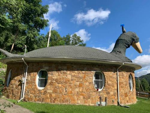 A round stone house with a whimsical duck-shaped roof and a blue object on its head, set against a blue sky.