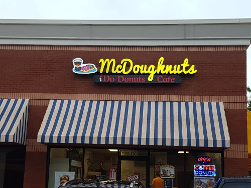Sign for "McDoughnuts i Do Donuts Cafe" with colorful graphics and striped awning. Open sign visible in the window.