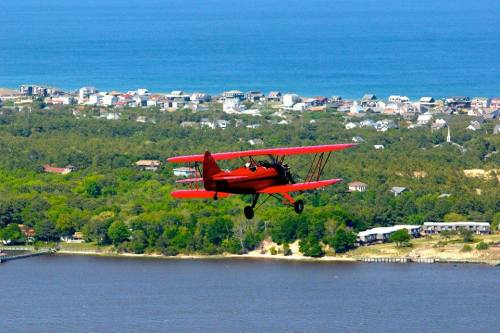 A red biplane flying over a coastal area with houses and trees, near a blue body of water.