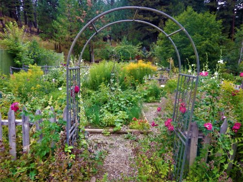 A garden archway leads into a lush, colorful garden filled with flowers and greenery, surrounded by trees.