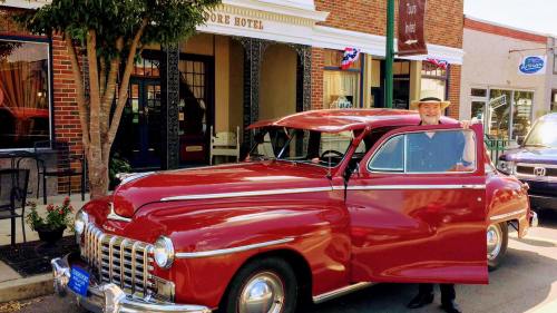 A man in a hat stands beside a vintage red car parked in front of a historic hotel on a sunny day.