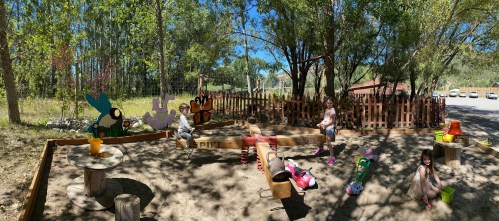 Children playing in a sunny sandbox area with colorful animal decorations and wooden play equipment surrounded by trees.