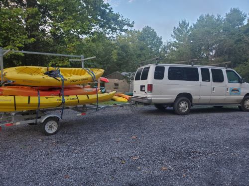 A white van towing a trailer loaded with yellow kayaks, parked in a gravel area surrounded by trees.