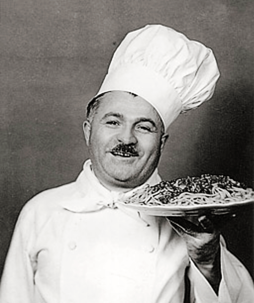 A smiling chef in a white uniform and tall hat holds a plate of spaghetti with sauce.