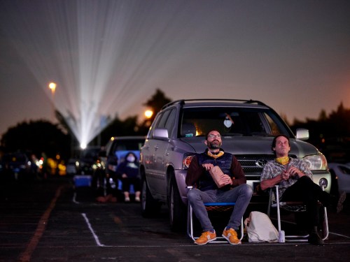 Two men sit in chairs outside their car at a drive-in movie, with a projector beam shining in the background.