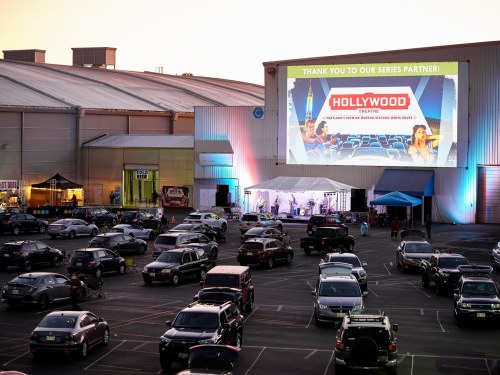 A drive-in event with cars parked, a large screen displaying "Thank You to Our Series Partner" and a live band performing.