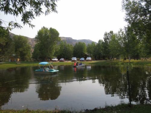 Two boats on a calm pond surrounded by green trees and parked cars in the background. Mountains in the distance.