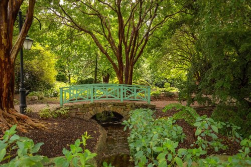 A serene garden scene featuring a turquoise bridge over a small stream, surrounded by lush greenery and trees.