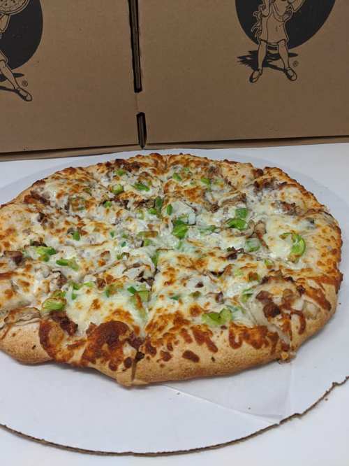 A freshly baked pizza topped with cheese, green peppers, and chunks of meat, placed on a white surface near pizza boxes.