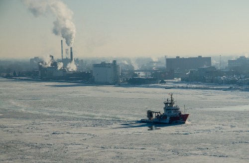 A ship navigates through icy waters near industrial buildings emitting smoke in a winter landscape.