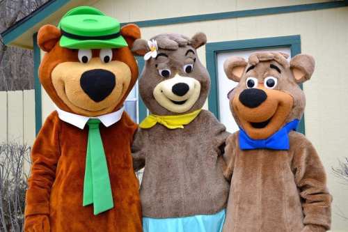 Three cartoon bear characters in costumes, smiling together outside a building. One wears a green hat and tie.