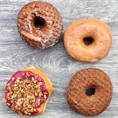 Four assorted donuts on a wooden surface: glazed, plain, and two with toppings, including one with granola and berry icing.