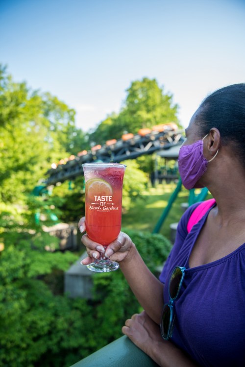 A woman in a purple shirt holds a colorful drink with a lemon slice, wearing a mask, with a roller coaster in the background.